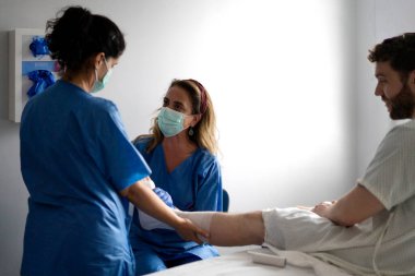 Hastane odasında erkek bir hastanın bacağını saran yüz maskesi takan iki hemşire.