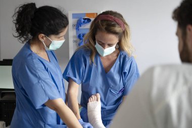 Yüz maskesi takan iki kadın doktor kırık bilekli bir hastayla ilgileniyor.