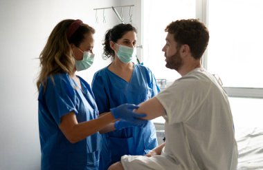 Ameliyat maskesi takan iki hemşire hastane odasında erkek bir hastanın kolunu inceliyor.