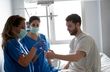 Maske ve eldiven takan iki hemşire hastane yatağında oturan bir erkek hasta için gazlı bez hazırlıyor.