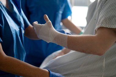 Hemşire, hastane yatağında iyileşen hastanın bandajlı elini inceliyor.