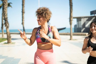 Güneşli bir günde sahilde koşan iki kadın biri bir şeyi işaret ederken...