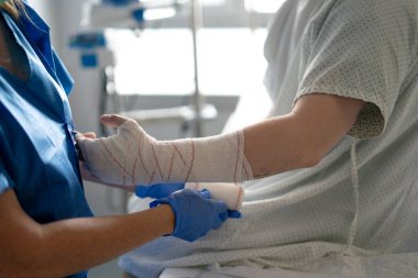 Hemşire tıbbi bakım sağlıyor, hastane odasında yaralı bir hastanın elini bandajlıyor, sağlık desteği ve iyileşme gösteriyor.