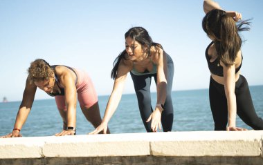 Deniz kenarındaki beton duvarda yoga yapan üç kadın.