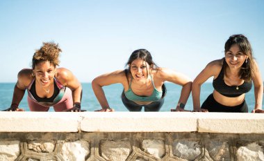 Üç kadın deniz kenarındaki taş bir duvarda şınav çekiyorlar. Açık hava egzersizinin tadını çıkarıyorlar.