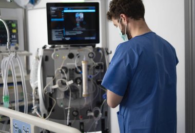 Hastane odasındaki diyaliz makinesinde cerrahi maske takan doktor hasta durumunu izliyor.