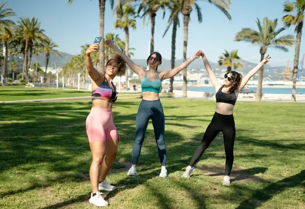Üç bayan sporcu, palmiye ağaçlarıyla dolu bir parkta antrenman yaptıktan sonra selfie çekiyorlar.