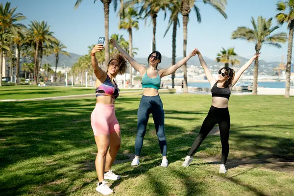 Üç bayan sporcu, palmiye ağaçlarıyla dolu bir parkta antrenman yaptıktan sonra selfie çekiyorlar.