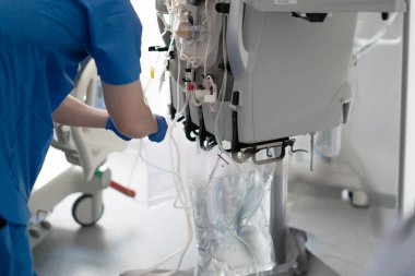 Hemşire mavi önlük ve eldiven giyip hastane odasındaki serum torbasına diyaliz makinesini bağlıyor.