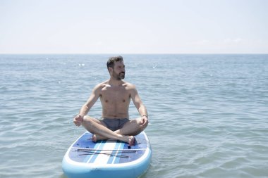 Rahat bir adam yoga yapıyor ve lotus pozisyonunda meditasyon yapıyor. Kürek tahtasında, okyanusun huzurunun tadını çıkarıyor.