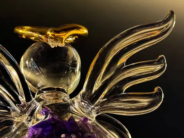 Koyu arkaplanda hafif ışıklandırmayla vurgulanmış, altın bir halesi ve mor bir çiçek detayı olan cam melek heykelciğinin yakın görüntüsü..