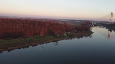 Ufuk çizgisine doğru uçan nefes kesici bir insansız hava aracı, ormanlarla çevrili bir nehre doğru uçuyor ve günbatımında büyük bir kablo destekli köprü var.