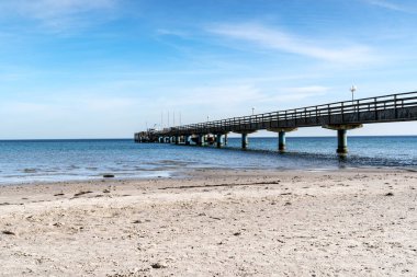 Uzakta mavi gökyüzü olan bir iskele görülebilir. Baltık Denizi sahili boş ve su sakin.