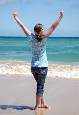 Bir kadın kumsalda kolları havada duruyor. Plaj, arkasında okyanus olan güzel bir mavi renktir. Kadın mutlu ve anın tadını çıkarıyor.