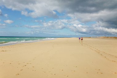 İspanya 'daki Fuerteventura kanarya adalarında iki kişi sahilde yürüyor. Kumsal kumlu ve arka planda bir su kütlesi var.