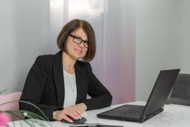 Bir kadın ofiste dizüstü bilgisayarı ve faresi olan bir masada oturuyor. Gözlük ve siyah bir ceket giyiyor.