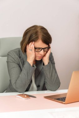 Bir kadın bir dizüstü bilgisayar ve bir fareyle masada oturuyor. Gözlük takıyor ve kafası elinde.