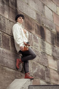 Beyaz kazak ve siyah pantolon giyen bir kadın duvara yaslanıyor. Elinde bir kamera tutuyor.