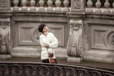 Siyah şapkalı ve beyaz kazaklı bir kadın taş bir duvarın önünde duruyor. Elinde bir kamera tutuyor.