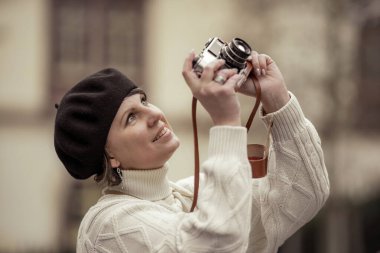 Siyah şapkalı ve beyaz kazaklı bir kadın fotoğraf çekiyor. Taş bir duvarda duruyor.