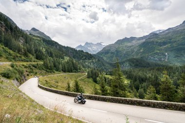 İsviçre Alp Dağları 'nda bir motosiklet dolambaçlı bir yoldan gidiyor. Yol ağaçlarla çevrili ve gökyüzü açık.