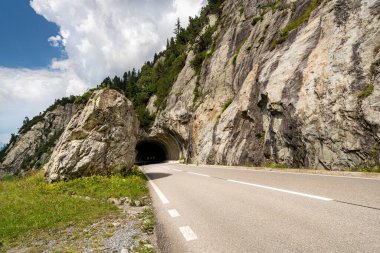 İsviçre Alplerinde ortasında bir tünel olan bir yol. Tünel kayalarla çevrili. Yol asfaltlı ve beyaz çizgileri var.
