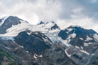 İsviçre Alpleri 'nde tepesinde kar ve buz olan bir dağ sırası. Dağlar yeşil çimlerle kaplıdır. Gökyüzü bulutlu ve gri