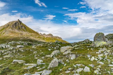 Kayalık bir arazinin arka planında bir dağ var. Gökyüzü bazı bulutlarla mavidir. Ön planda büyük bir kaya var.