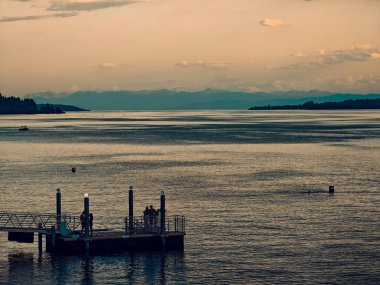 Denizde bir rıhtım ve üzerinde birkaç kişi oturuyor. Su sakin ve berrak.
