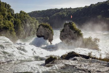 Schaffhausen İsviçre 'de bir Ren şelalesi. Havada kırmızı bir bayrak sallanıyor. Su iki büyük kayaya çarpıyor.