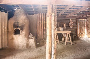 Eski bir ev. Oda toprakla dolu ve ortasında bir odun sobası var. Odada bir masa var ve sağ tarafta da bir kapı var. Oda kırsal bir kulübe ya da küçük bir atölye gibi görünüyor.