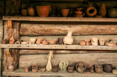 Eski bir ev. Oda toprakla dolu ve ortasında bir odun sobası var. Odada bir masa var ve sağ tarafta da bir kapı var. Oda kırsal bir kulübe ya da küçük bir atölye gibi görünüyor.