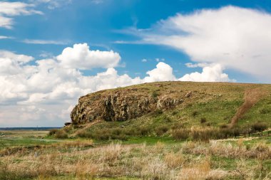Kayalık bir uçurumu ve çimenli bir arazisi olan bir tepe. Gökyüzü mavi ve bulutlu.