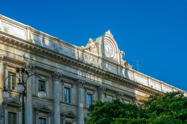 İtalya 'nın Milano şehrinde saati olan yüksek bir bina. Binanın birçok penceresi var ve taştan yapılmış. Binanın önünde ağaçlar var ve yakınlarda bir sokak lambası var.