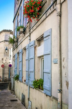 Konyak Fransa 'da bir bina. Mavi panjurları ve kırmızı çiçekleri var. Panjurlar açık. Binanın sol tarafında kırmızı bir tabela var.