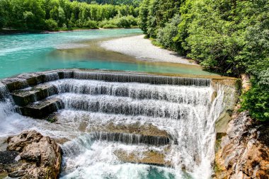 Altından nehirler akan bir şelale. Su berrak ve mavi. Suda ve yerde kayalar var.