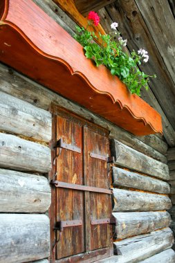 Ahşap pencereli ve tepesinde saksı olan bir kır evi. Saksı pembe bir çiçek içeriyor, bu da kabinin içine biraz renk katıyor. Pencere biraz aralık.