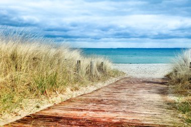 Almanya 'da Baltık Denizi' ne giden kırmızı tahta bir tahta yol. Tahta yol uzun otlarla çevrilidir. Kumsal güzel mavi bir renktir.