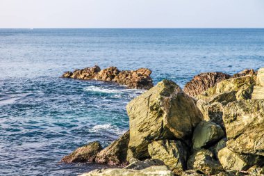 Önünde büyük bir kaya olan kayalık bir kıyı şeridi. Su sakin ve gökyüzü berrak.