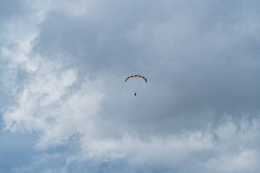 Gökyüzünde bir paraşüt uçuyor. Paraşüt turuncu ve sarıdır. Gökyüzü bulutlu.