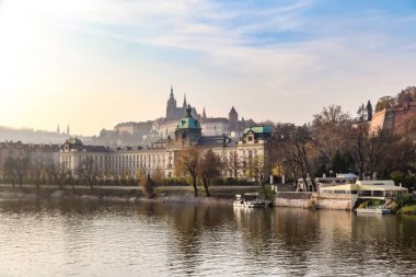 Prag Çek Cumhuriyeti 'nin içinden bir nehir akan ve arkasında büyük bir bina bulunan bir şehri. Gökyüzü bulutlu ve su sakin.