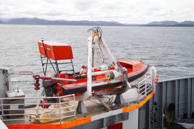 Kırmızı bir üstü ve beyaz bir tabanı olan bir tekne suda yüzüyor. Tekne turuncu ve beyaz. Kurtarma botu. Tekne su kenarına yakın bir yerde ve etrafı dalgalarla çevrili.