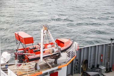 Kırmızı bir üstü ve beyaz bir tabanı olan bir tekne suda yüzüyor. Tekne turuncu ve beyaz. Kurtarma botu. Tekne su kenarına yakın bir yerde ve etrafı dalgalarla çevrili.