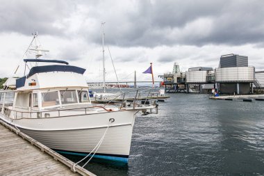 Norveç 'in Stavanger kentindeki Norveç Petrol Müzesi' nin arka planında bulutlu bir gökyüzü bulunan bir marinaya bir tekne yanaştı. Tekne beyaz ve mavi ve bir rıhtıma bağlı. Sahnenin sakin ve huzurlu bir havası var.