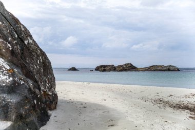 Norveç 'te kayalık bir plaj ve önünde büyük bir kaya var. Gökyüzü bulutlu ve su mavi.
