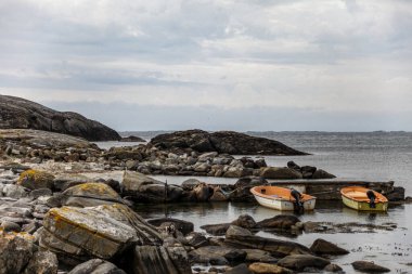 Norveç 'te kayalık bir kıyıda iki küçük tekne suda duruyor. Su sakin ve gökyüzü bulutlu. Sahnenin huzurlu ve huzurlu bir atmosferi var.