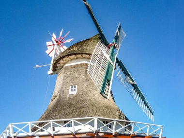 Beyaz çatılı ve kırmızı kapaklı bir yel değirmeni. Yel değirmeni uzun ve çok detay içeriyor. Gökyüzü mavi ve berrak