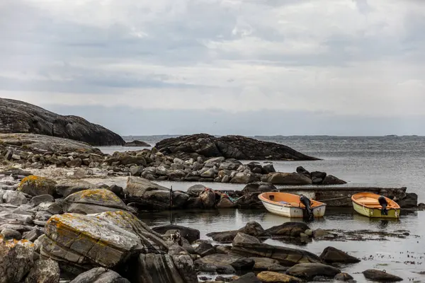 Norveç 'te kayalık bir kıyıda iki küçük tekne suda duruyor. Su sakin ve gökyüzü bulutlu. Sahnenin huzurlu ve huzurlu bir atmosferi var.