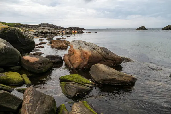 Norveç 'te kayalık bir kıyıda iki küçük tekne suda duruyor. Su sakin ve gökyüzü bulutlu. Sahnenin huzurlu ve huzurlu bir atmosferi var.