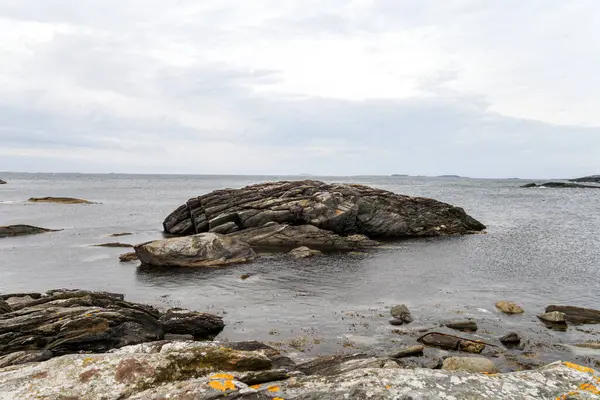 Norveç 'te kayalık bir plaj ve önünde büyük bir kaya var. Gökyüzü bulutlu ve su mavi.